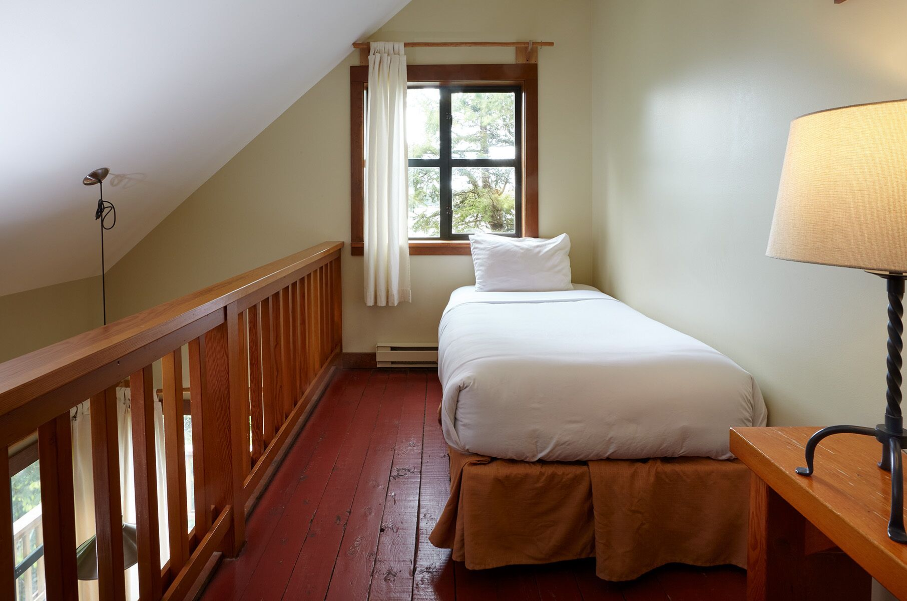 A bedroom with a single bed, a nightstand, a lamp, a window with curtains, and a wooden railing.