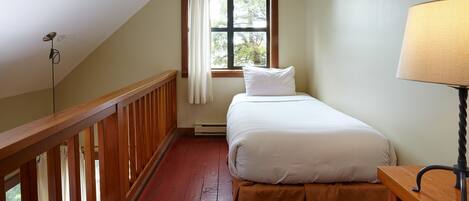 A bedroom with a single bed, a nightstand, a lamp, a window with curtains, and a wooden railing.