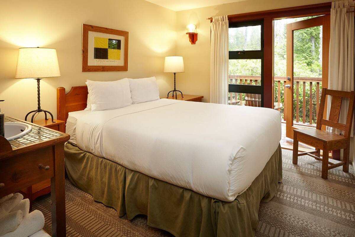 A hotel room with a large bed, two bedside tables with lamps, a wooden headboard, a framed artwork, a window with curtains, and a view of greenery outside.