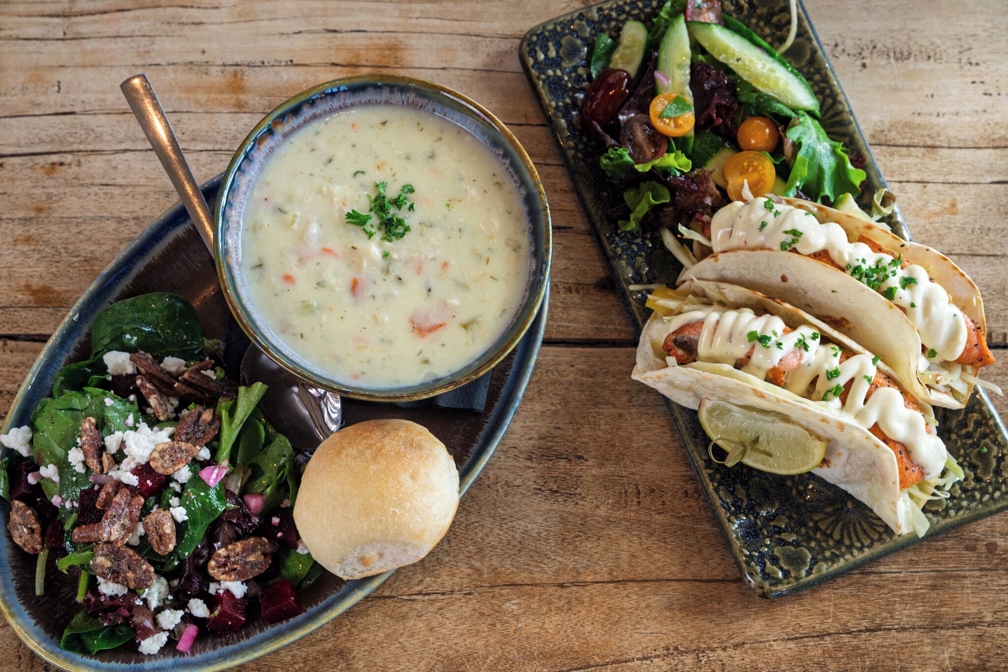 A bowl of creamy soup with a spoon, a plate of salad with nuts, and two wraps with meat and sauce.