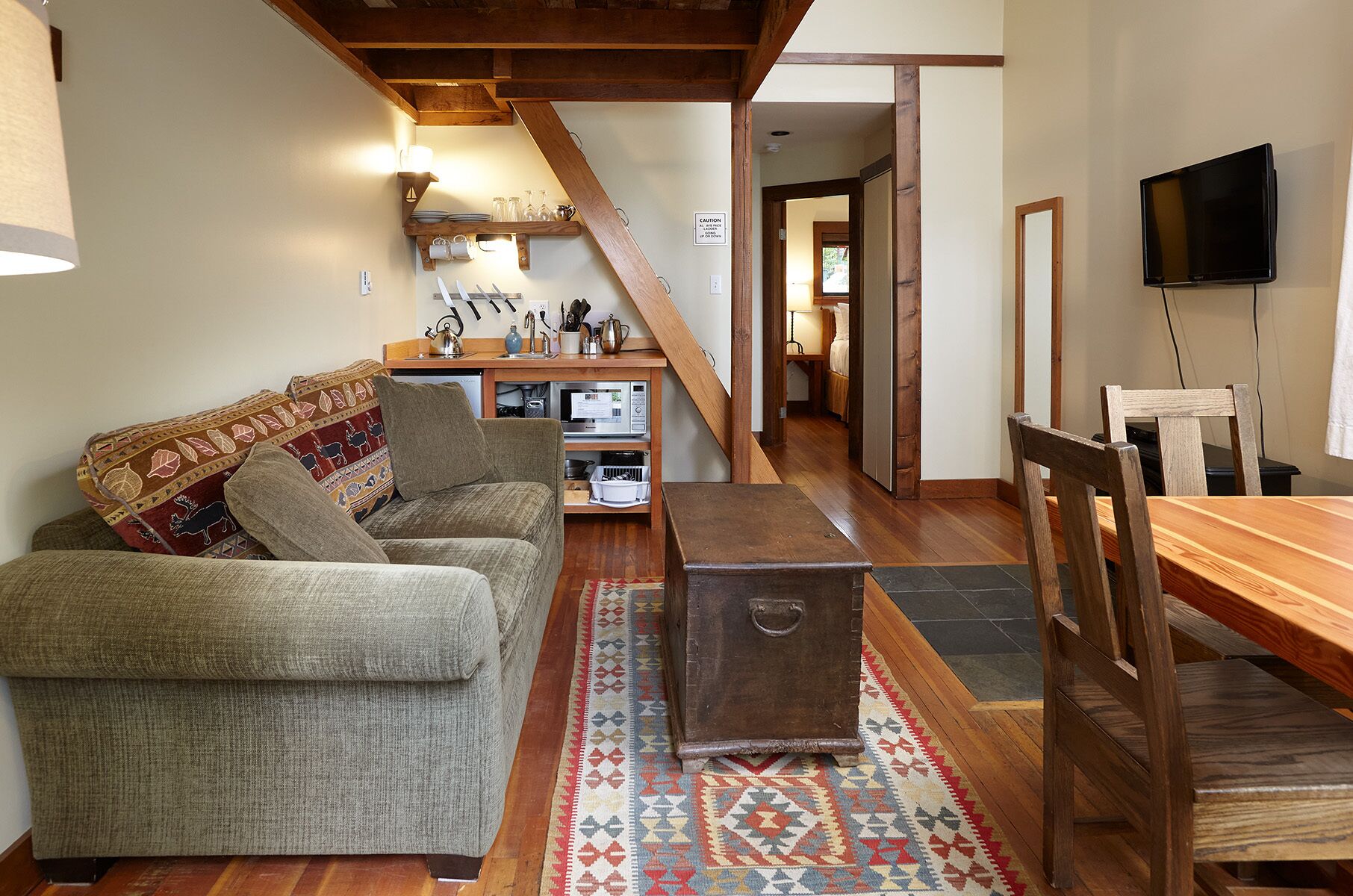 Una sala acogedora con un sofá, una mesa de centro de madera y un televisor. Se ve una escalera que conduce a otra habitación, y una zona de cocina con un lavabo y armarios.