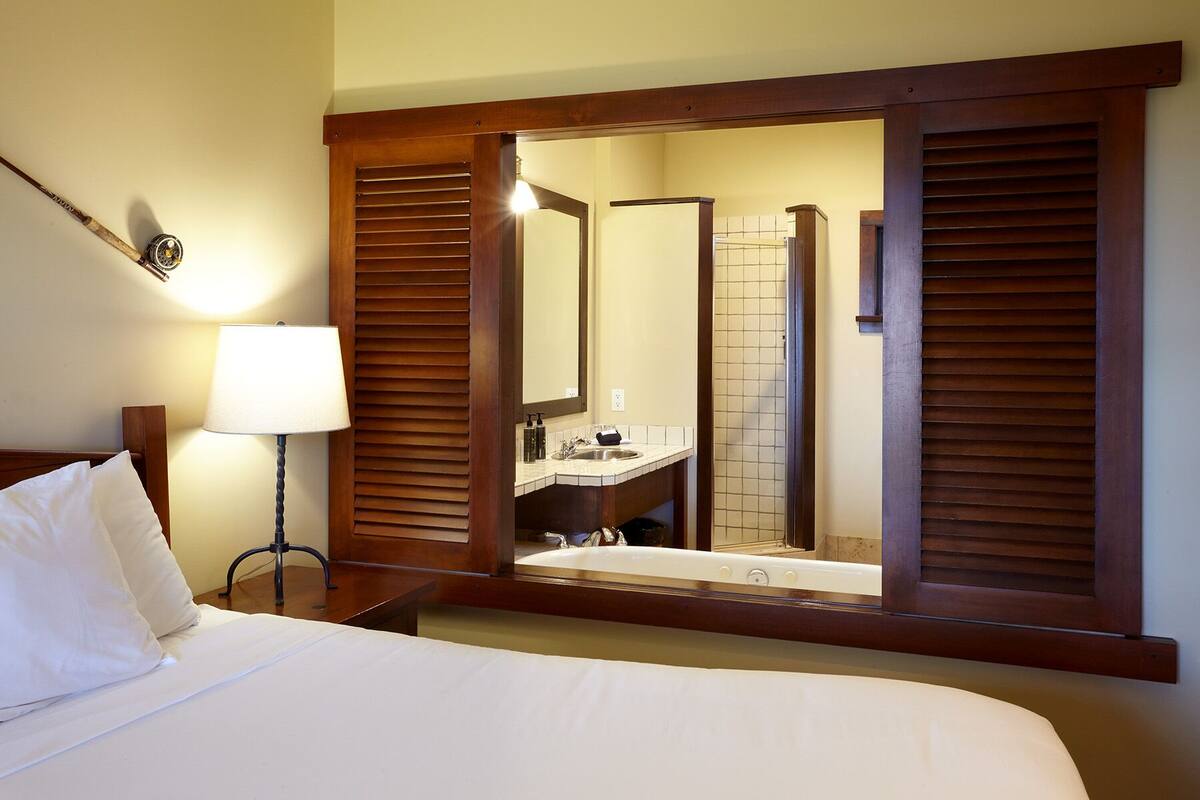 A hotel room with a bed, a bedside lamp, a wooden-framed mirror, and a bathroom visible through the mirror.