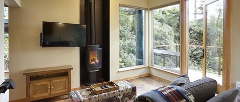 A cozy living room with a fireplace, a flat-screen TV, and large windows offering a view of the outdoors.