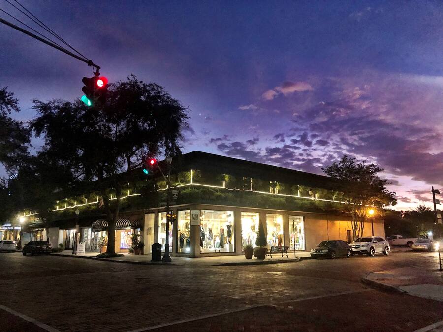 A building with multiple storefronts, a bench, and cars parked in front.