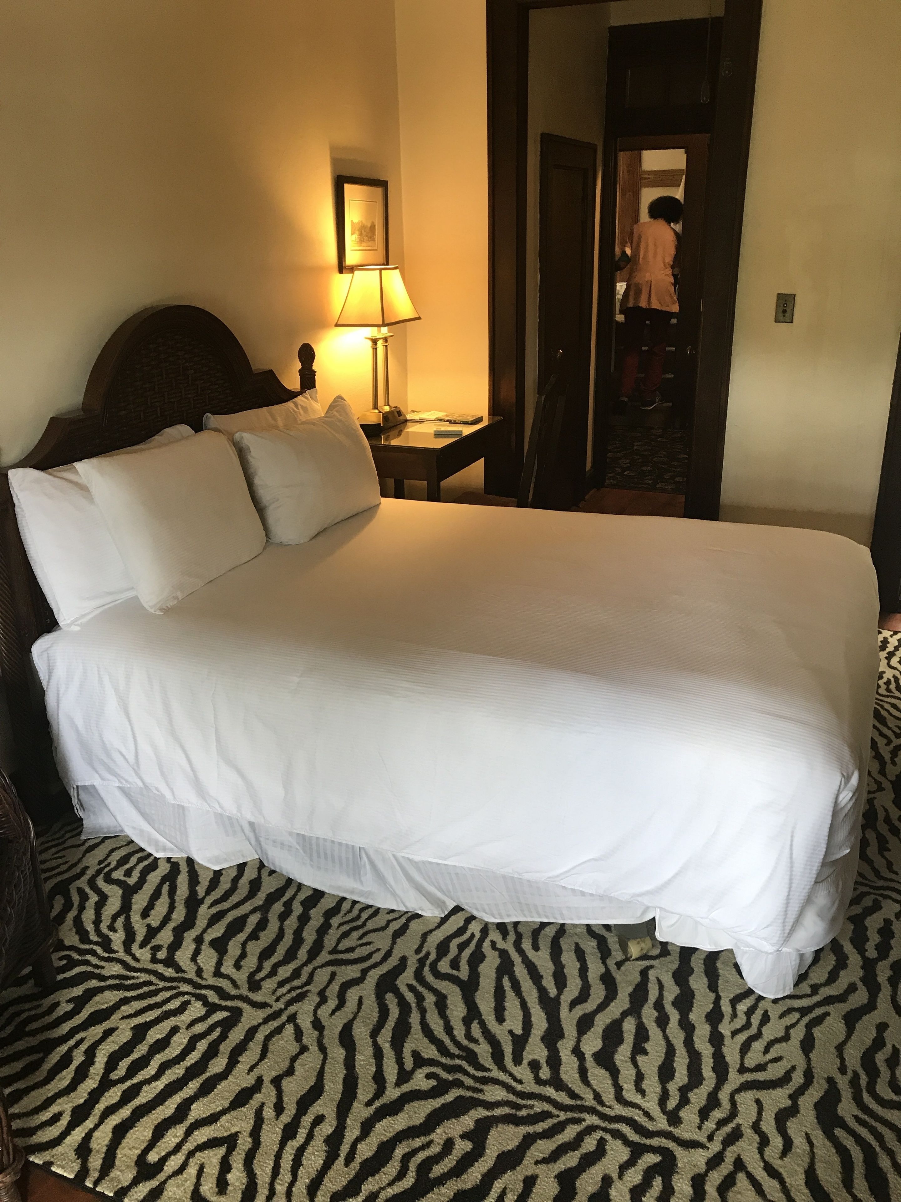A hotel room with a large bed, a nightstand with a lamp, a framed picture on the wall, and a zebra-patterned rug.