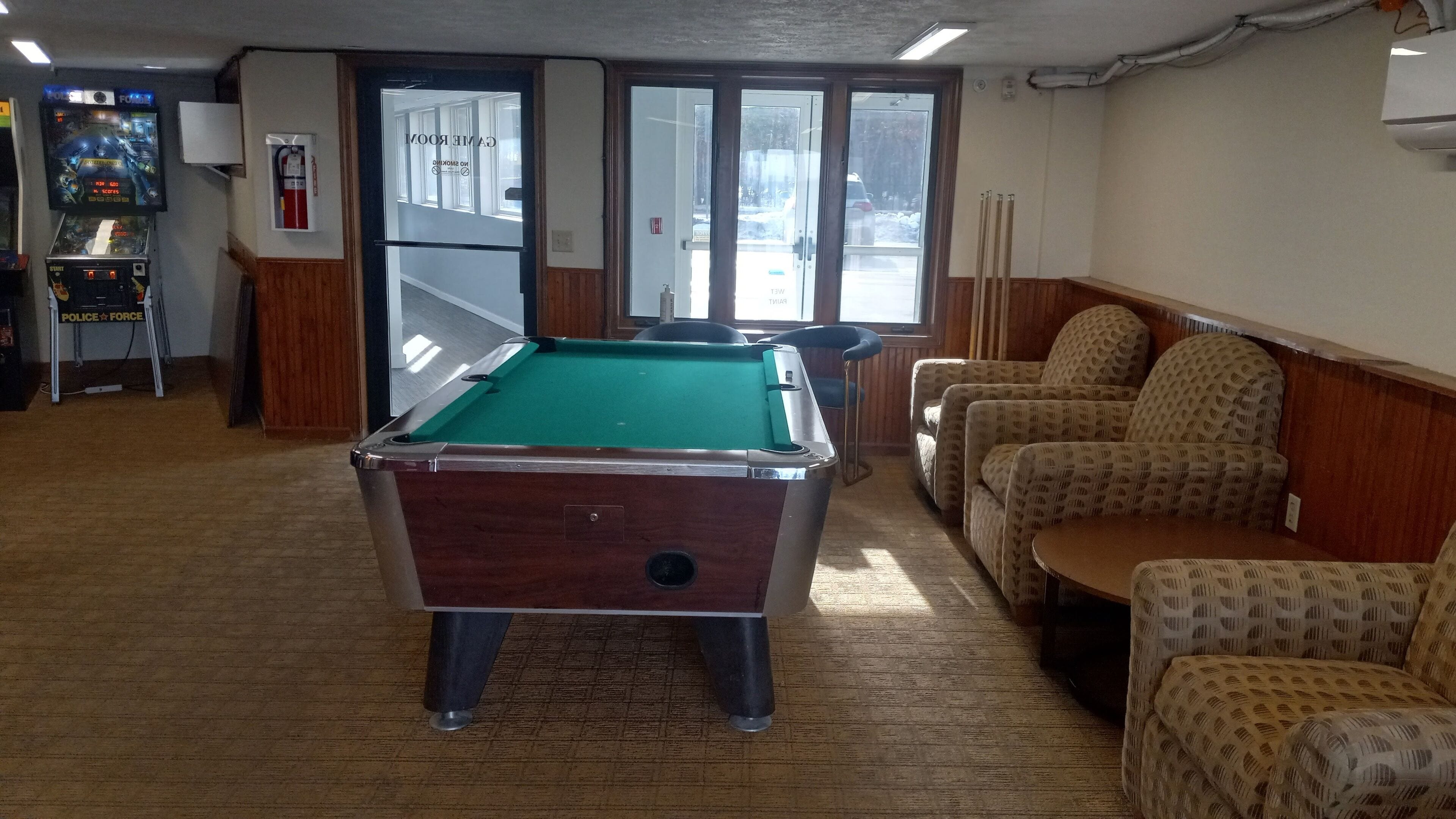 A pool table in a room with wooden flooring, a fire extinguisher, and a door leading outside. There are patterned chairs and a small table in the room.