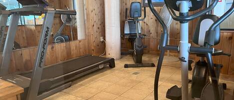 Ein Fitnessraum mit Laufband, Crosstrainer und Ergometer, Holzwänden und Fliesenboden.
