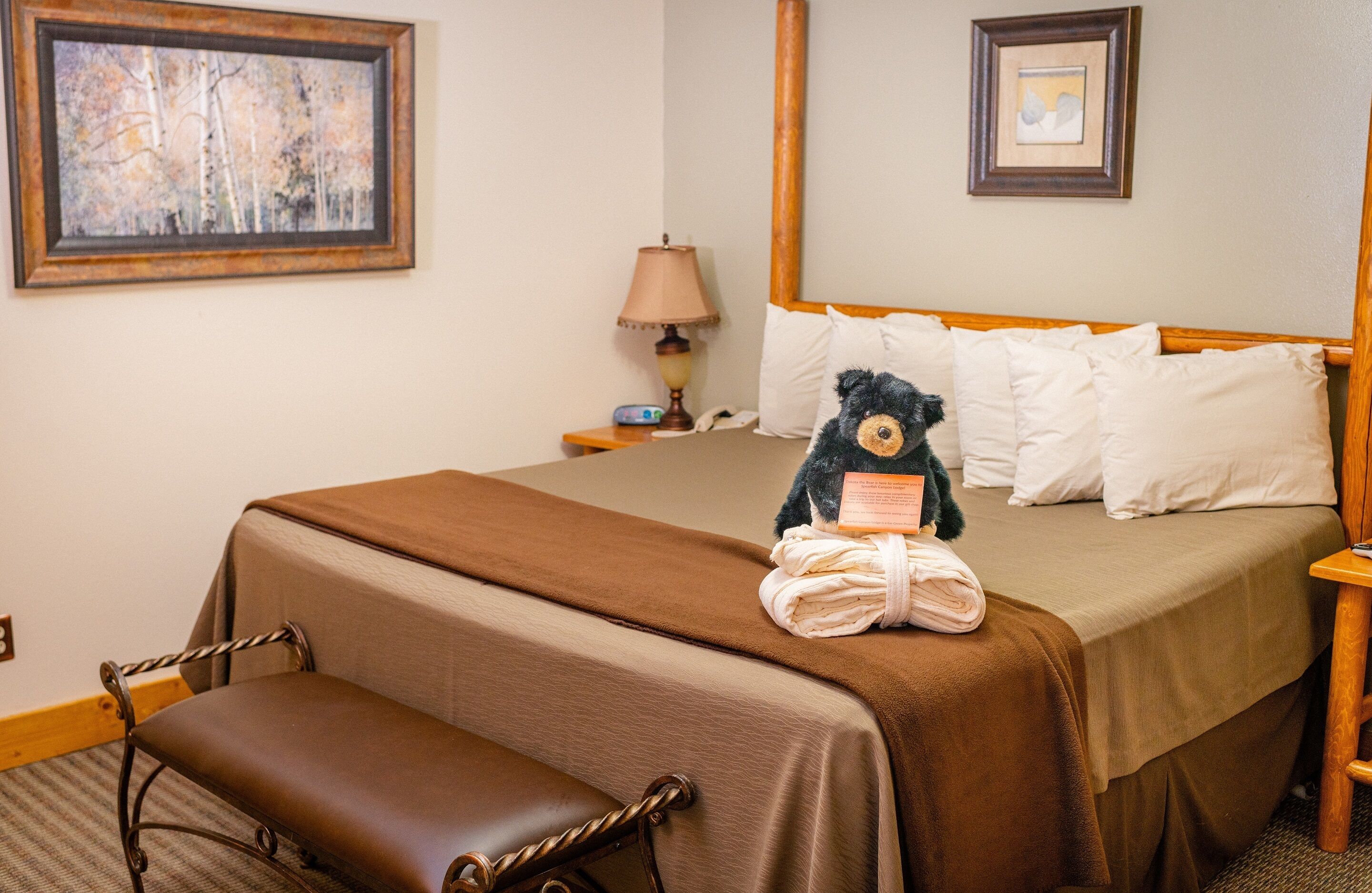 A neatly made bed with a brown bedspread, a bedside lamp, a framed picture on the wall, and a teddy bear on the bed.