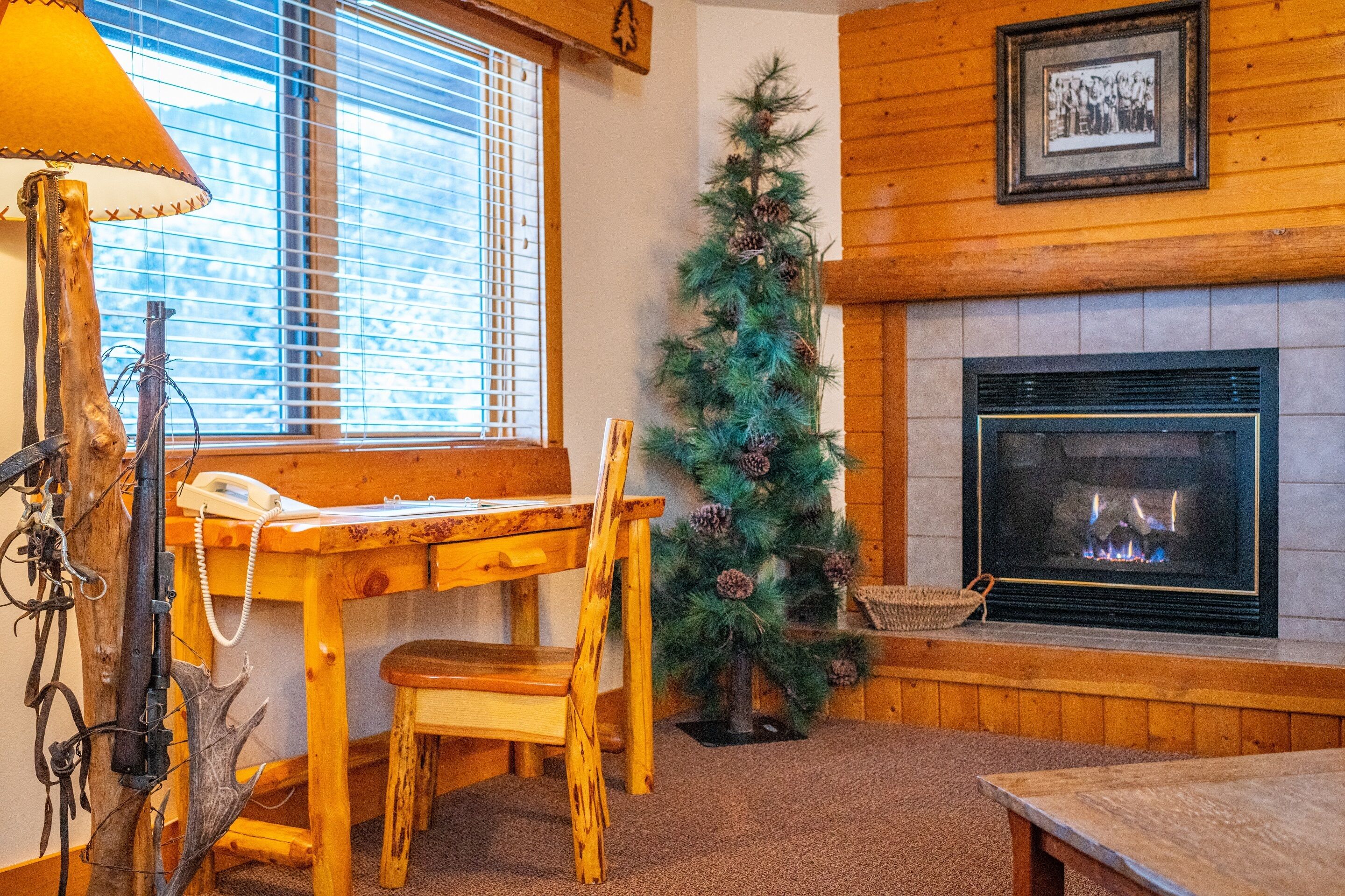A cozy room with a wooden table, a chair, a lamp, a fireplace, and a Christmas tree.
