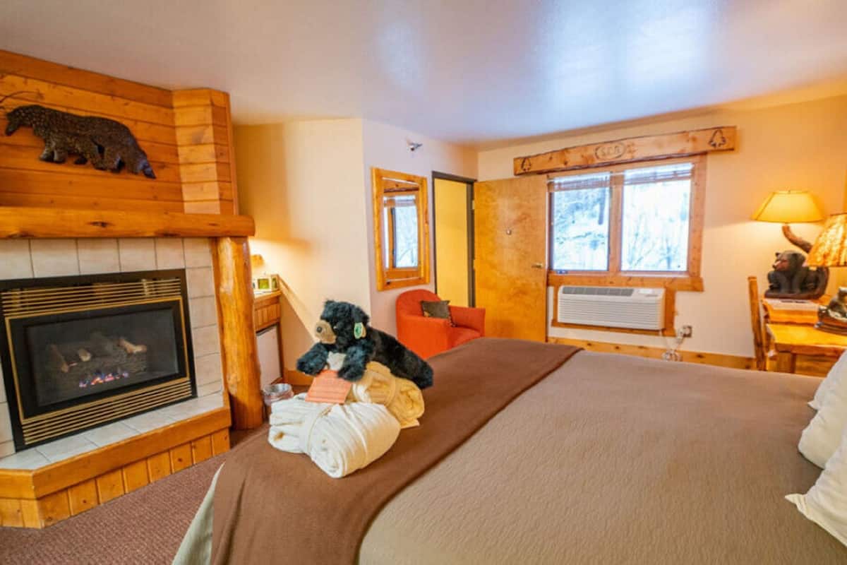 Una habitación acogedora con chimenea, una cama con una manta marrón, un peluche y una ventana con vistas a árboles.