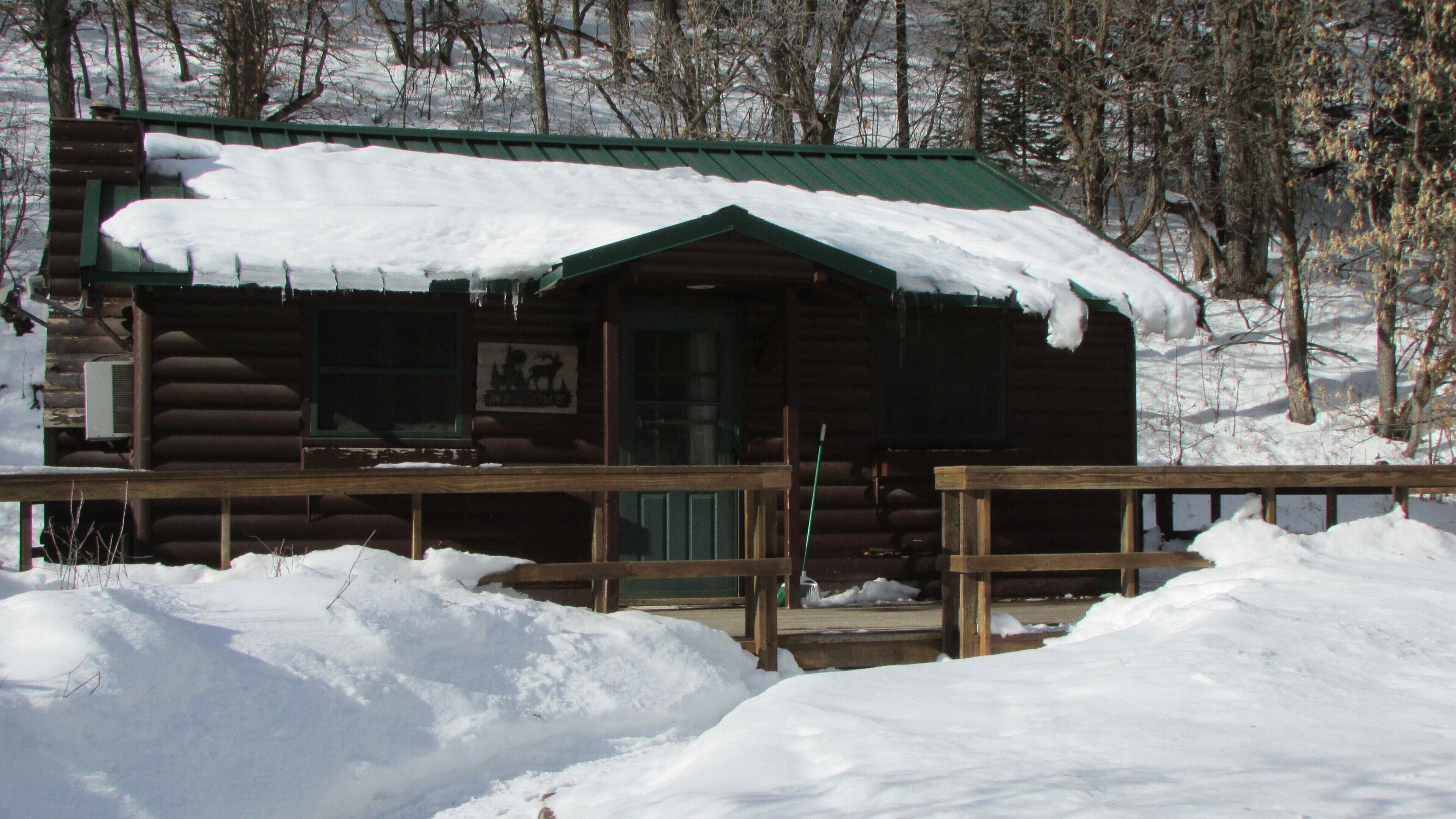 Una cabaña de troncos con techo verde y terraza de madera, rodeada de nieve.