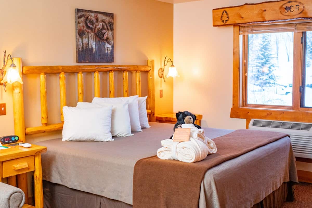 Un cuarto acogedor con cabecera de madera, cama con almohadas blancas, un osito de peluche y una ventana con vista a árboles.