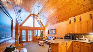 A spacious living area with a high ceiling, wooden beams, a fireplace, a large window, and a cozy seating area. - Spearfish Canyon Lodge (Lead)
