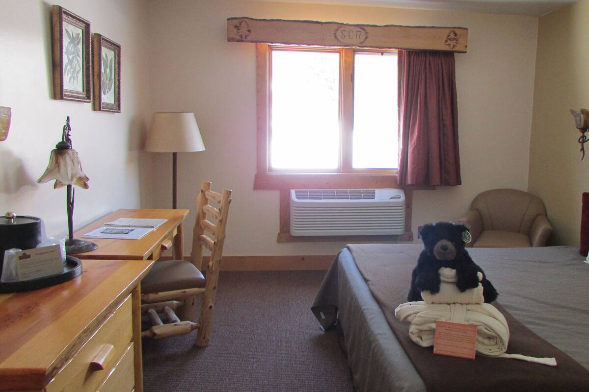 Una habitación de hotel con cama, escritorio con lámpara, silla, ventana con cortinas y aire acondicionado instalado en la pared.