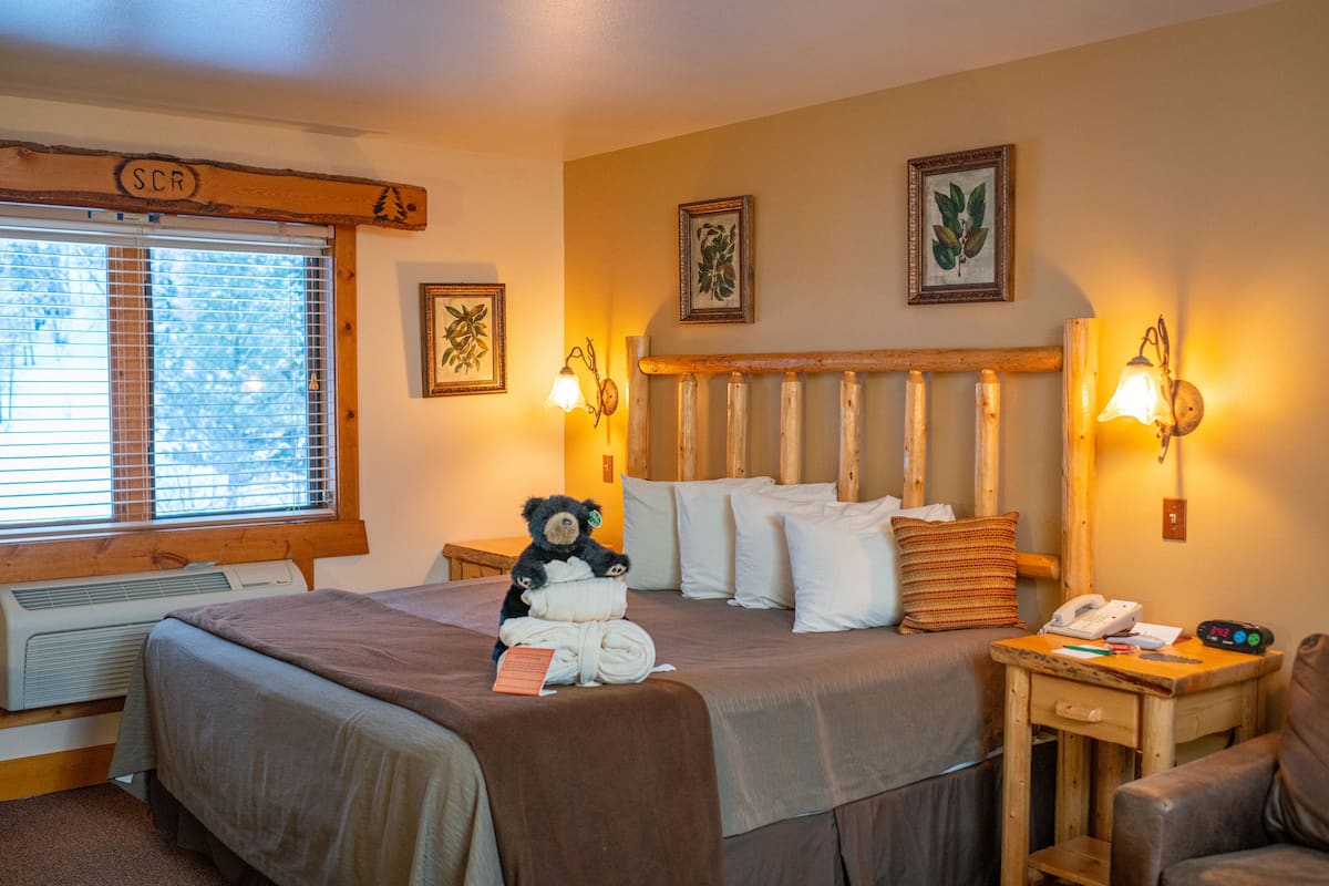Una habitación de hotel con una cama grande, una mesita de noche con lámpara, una silla y una ventana con persianas.