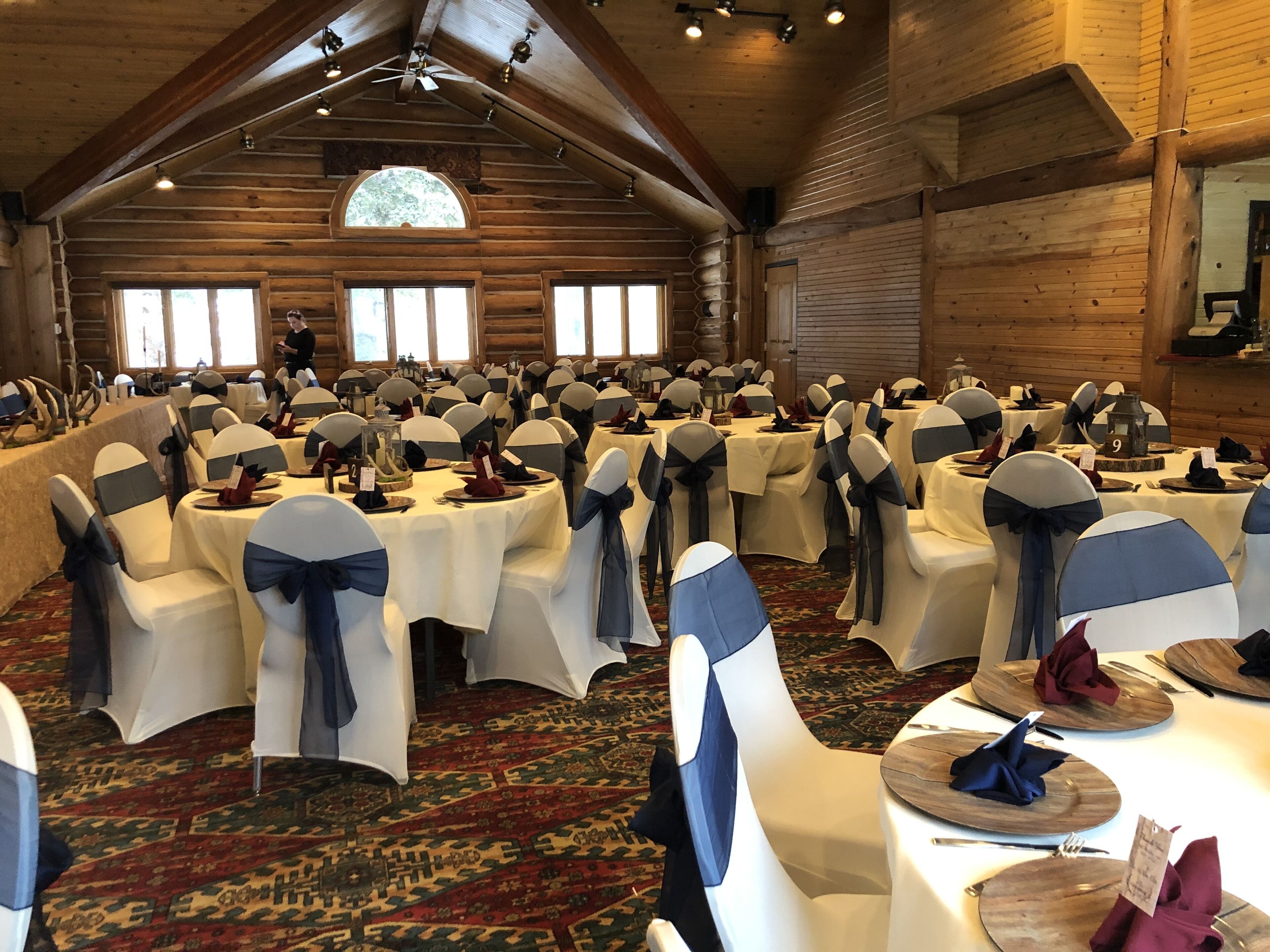 Une salle de banquet avec des tables rondes, des rubans de chaise blancs et bleus, et un décor intérieur en bois.