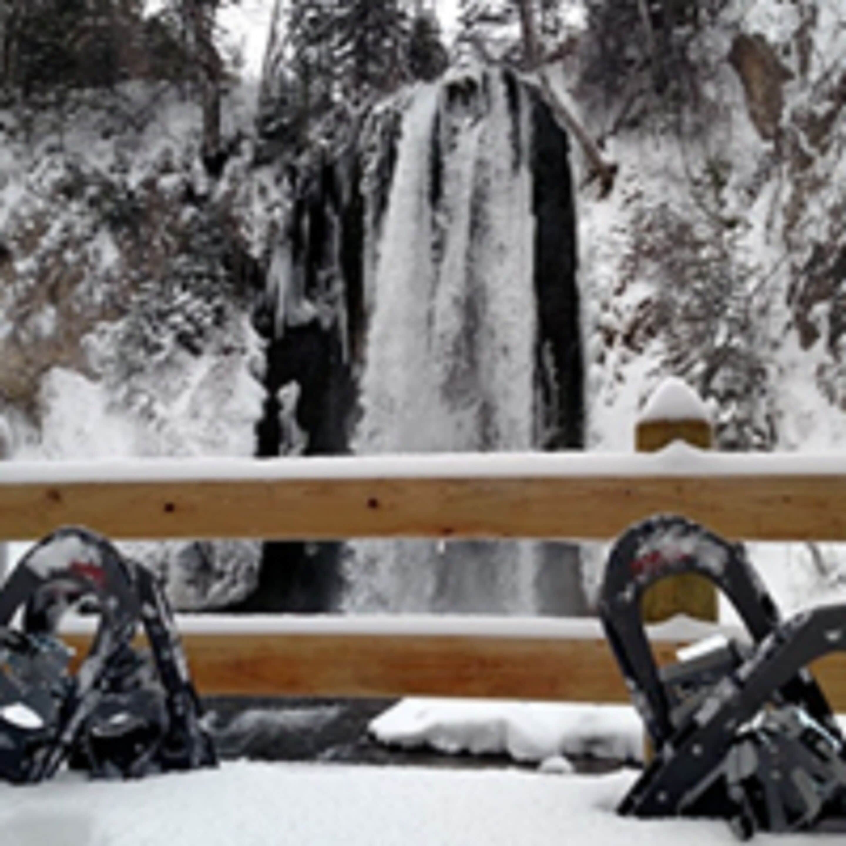 A wooden bench with snow on it, in front of a waterfall.