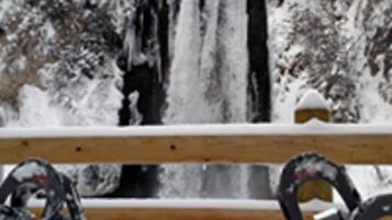 A wooden bench with snow on it, in front of a waterfall.