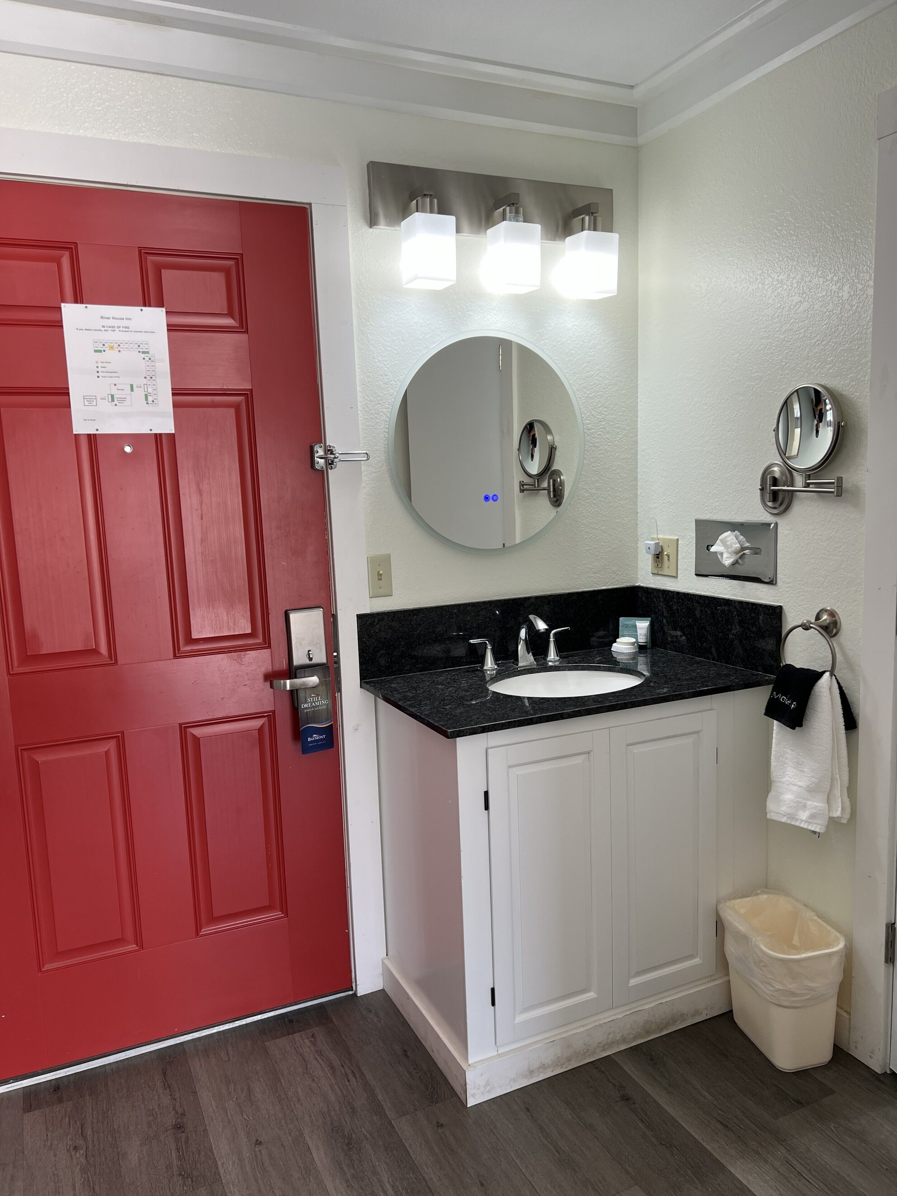 A bathroom with a red door, white cabinets, a black countertop sink, a mirror, and a trash can.
