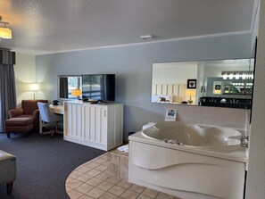 A hotel room with a desk, chair, television, large mirror, and a bathtub.