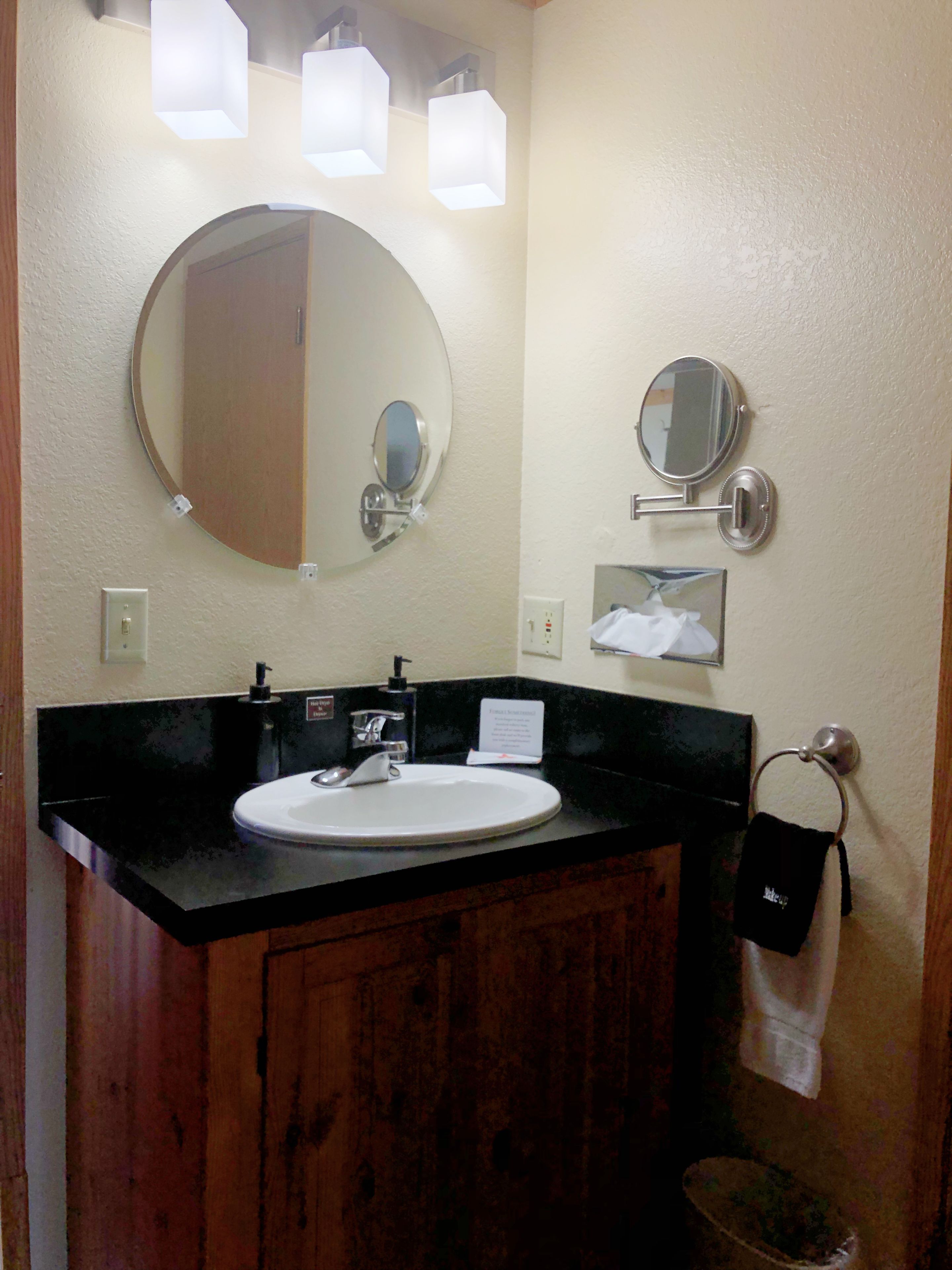 A bathroom with a round mirror, a rectangular mirror, and a sink with a black countertop. There are three wall-mounted lights above the mirrors.