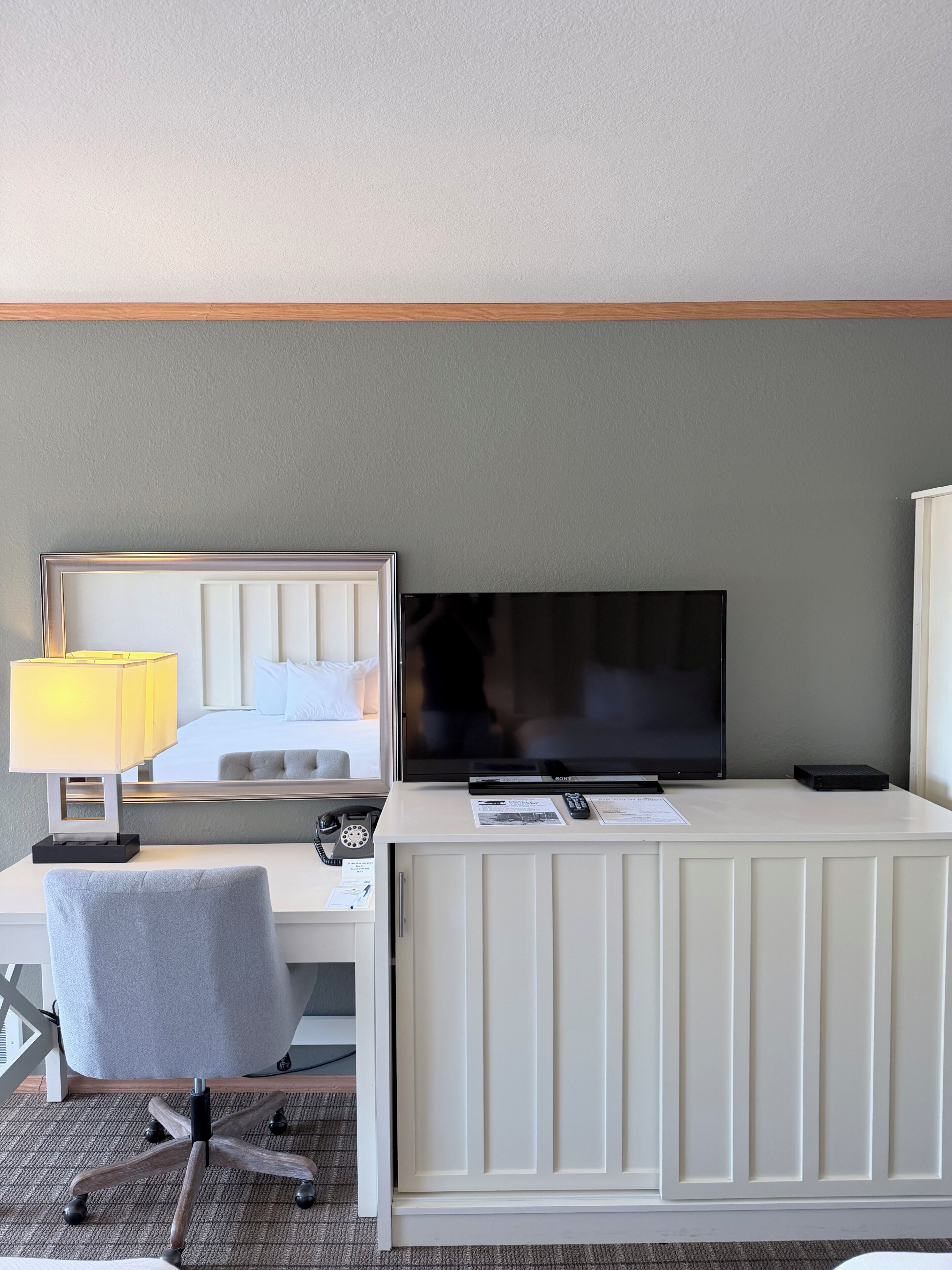 Quarto de hotel com cama, escrivaninha com luminária, televisão e uma cadeira.