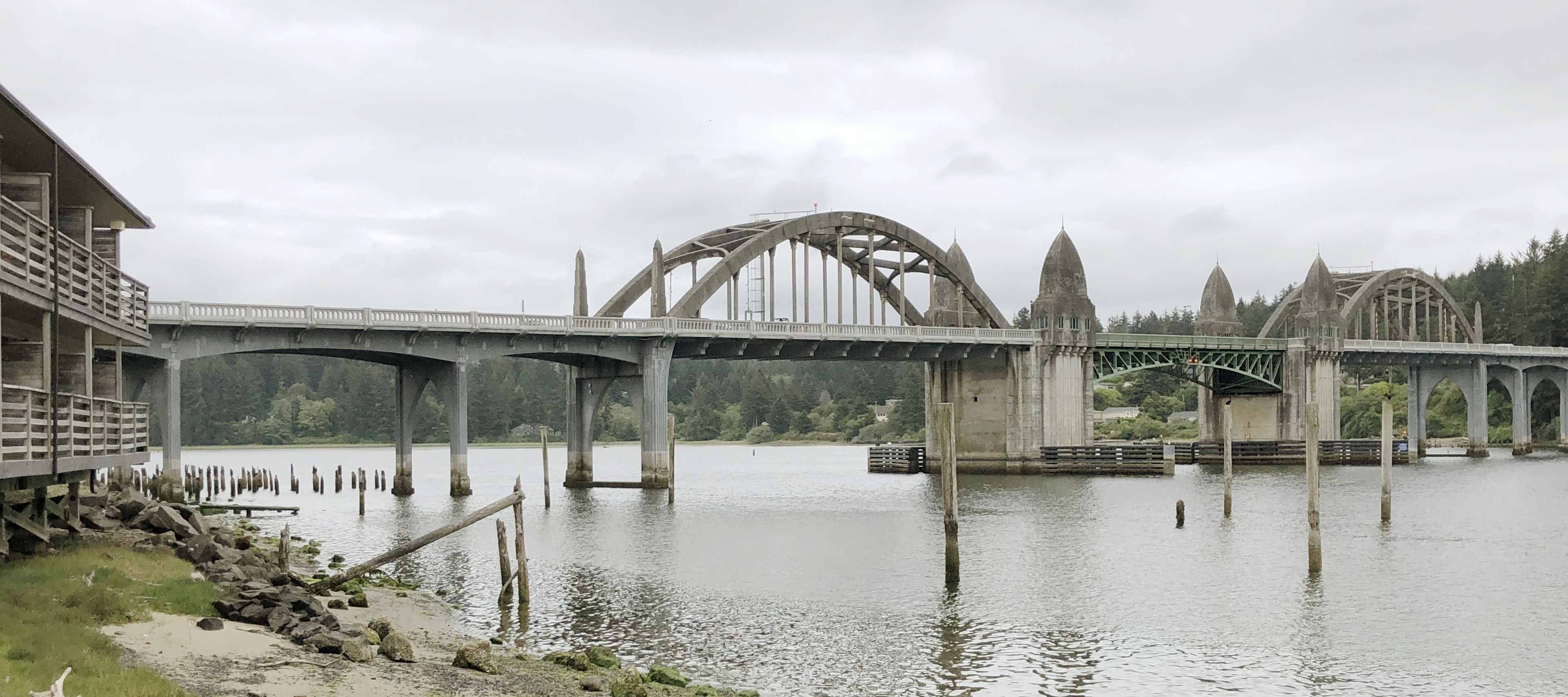 A large stone bridge with multiple arches over a calm river, surrounded by greenery and a small building with a balcony.