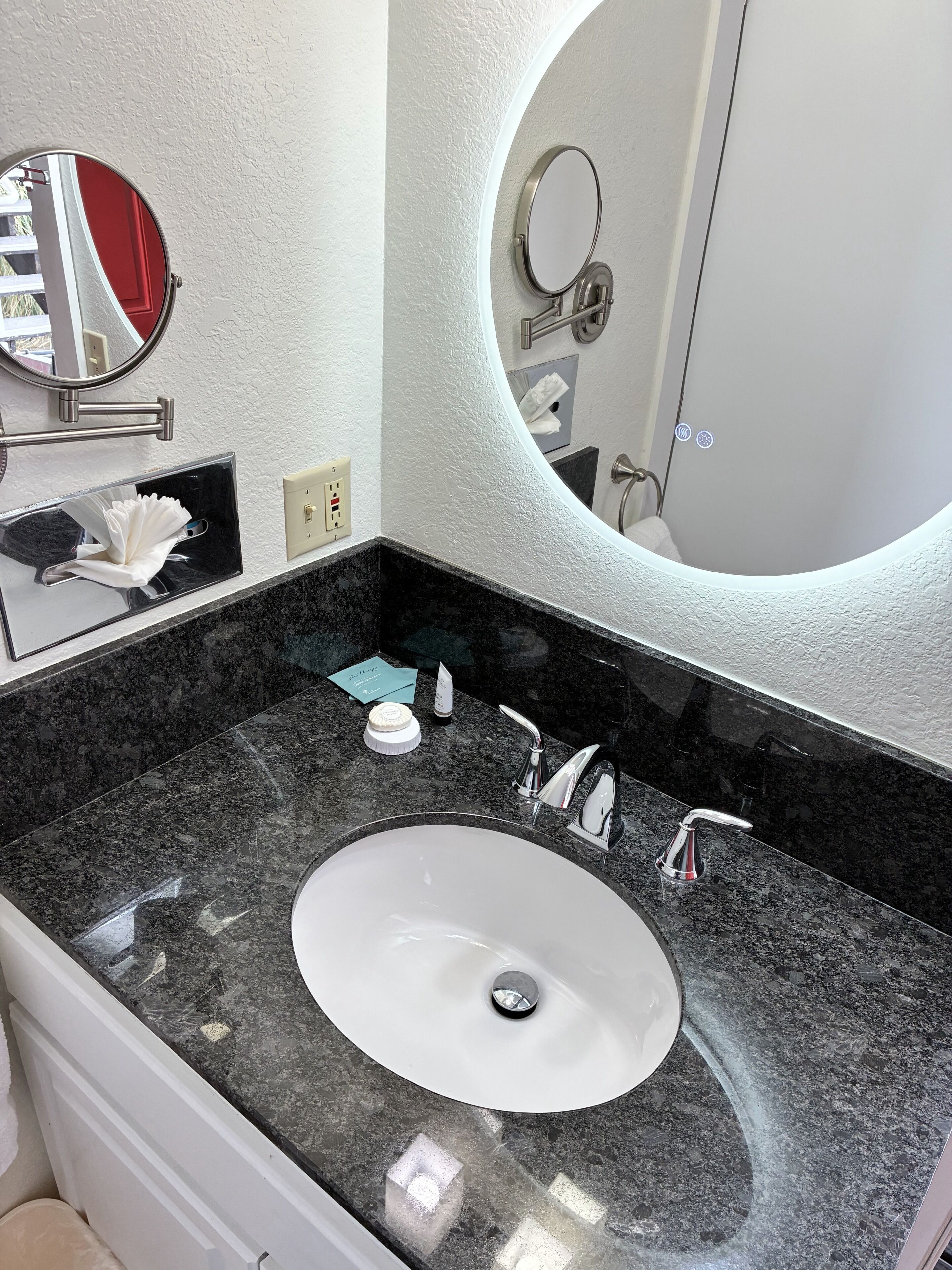A bathroom with a white sink, black granite countertop, two faucets, a mirror, and a towel dispenser.