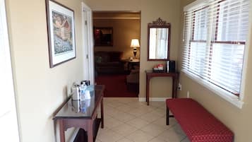 Habitación de hotel con una mesa de vidrio, un banco rojo, un cuadro enmarco, una lámpara y una ventana con persianas.