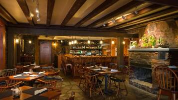 A restaurant with wooden tables and chairs, a bar area with shelves of bottles, and a stone fireplace.