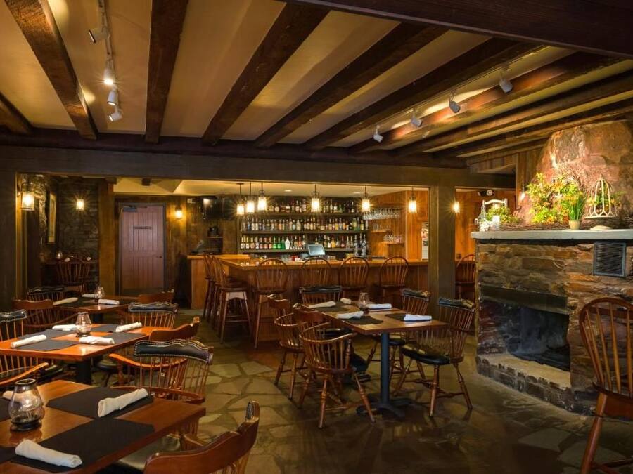 A restaurant with wooden tables and chairs, a bar area with shelves of bottles, and a stone fireplace.