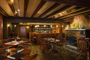 A restaurant with wooden tables and chairs, a bar area with shelves of bottles, and a stone fireplace.