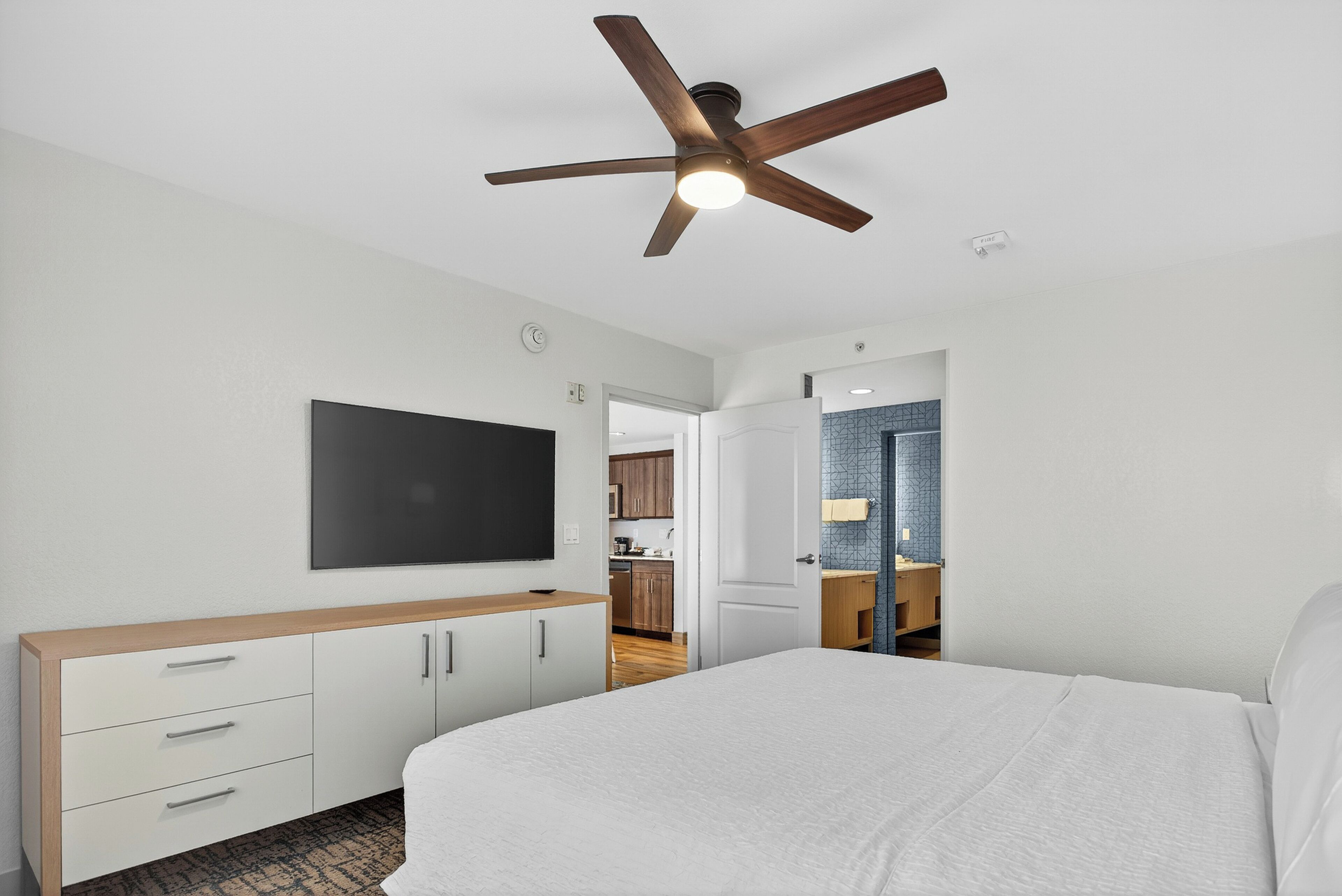 A modern hotel room with a large bed, a TV mounted on the wall, a wooden bedside table, and a ceiling fan.