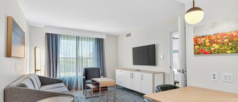 Ein modernes Hotelzimmer mit Sofa, einem kleinen Tisch, einem Esstisch, einem Fernseher und einem floralen Gemälde an der Wand.
