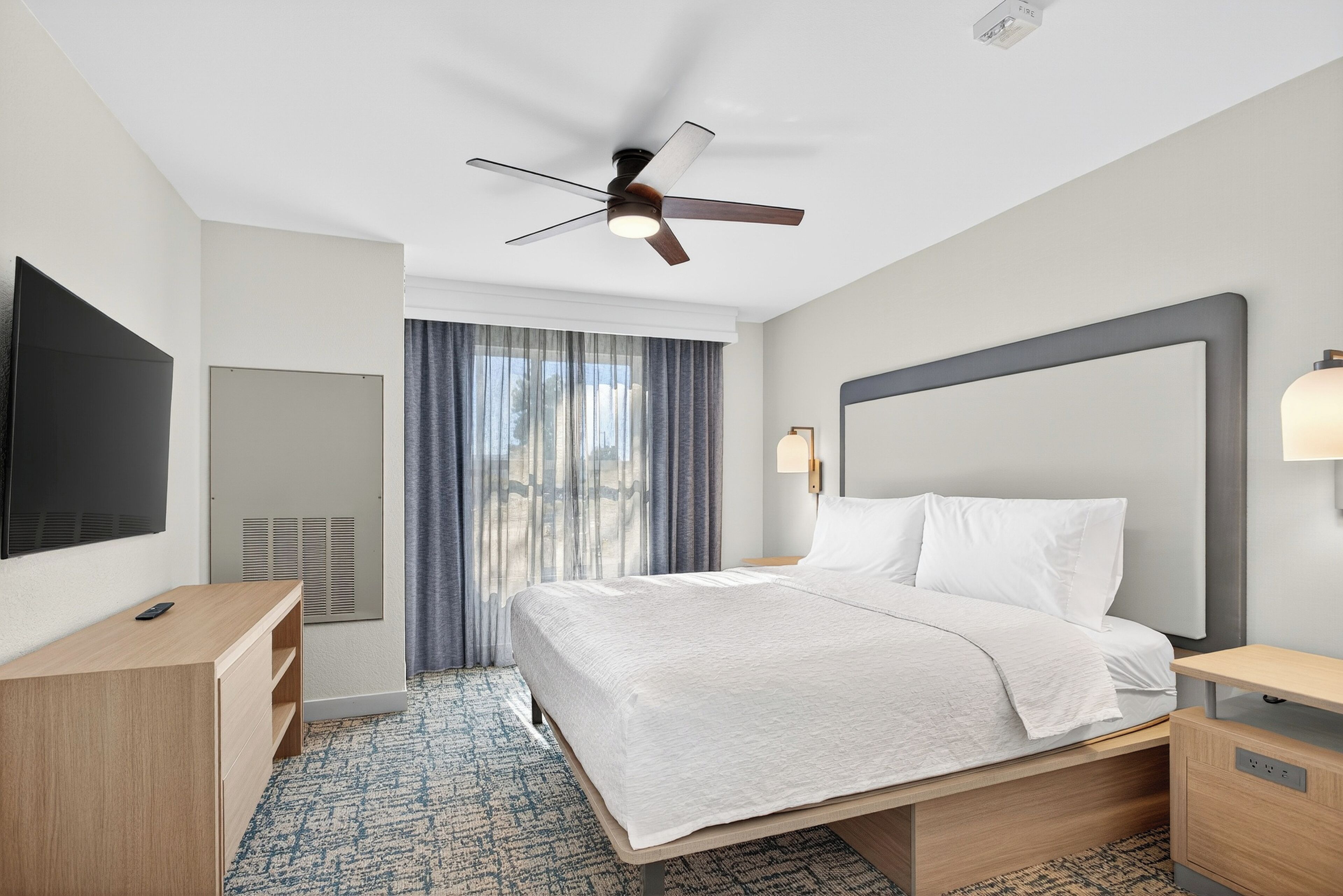 A hotel room with a bed, a desk, a television, a ceiling fan, and a window with curtains.