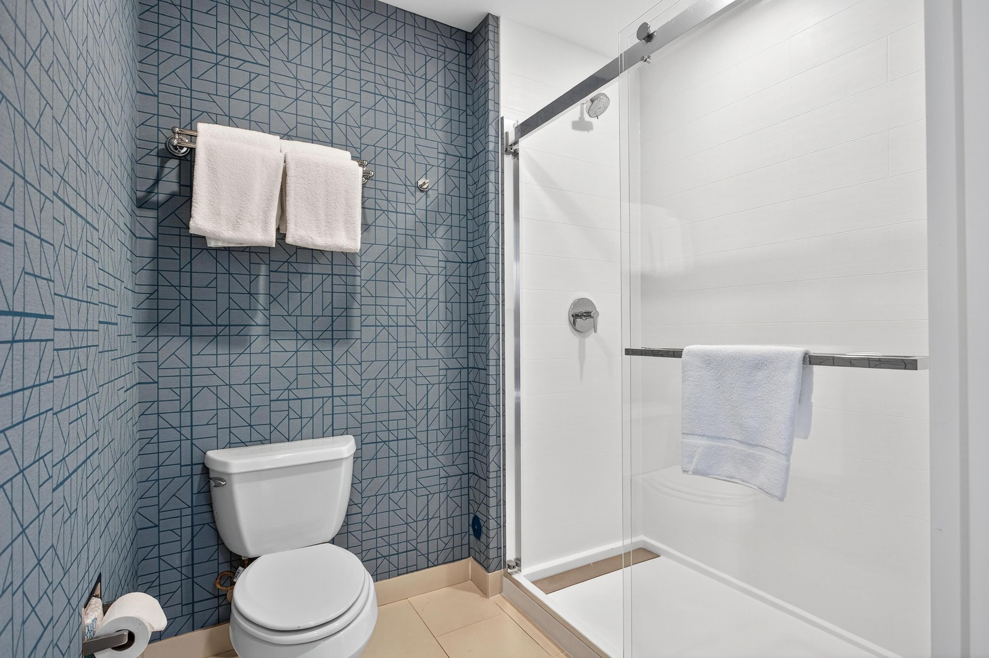 Ein Badezimmer mit geometrischen Fliesen an der Rückwand, einer Toilette, einer Duschkabine und einem Handtuchhalter mit zwei Handtüchern.