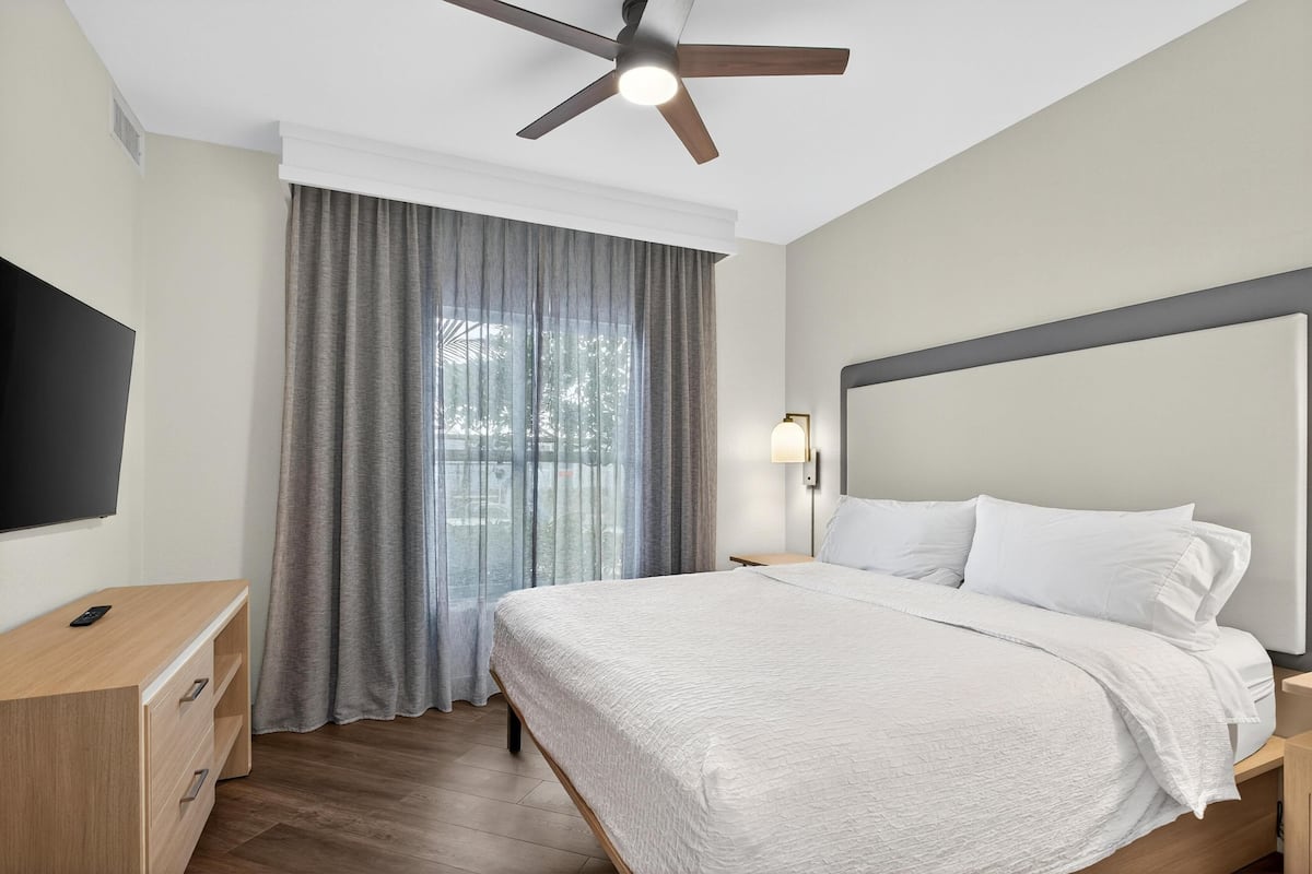 A hotel room with a bed, a desk, a ceiling fan, a window with curtains, and a wall-mounted TV.