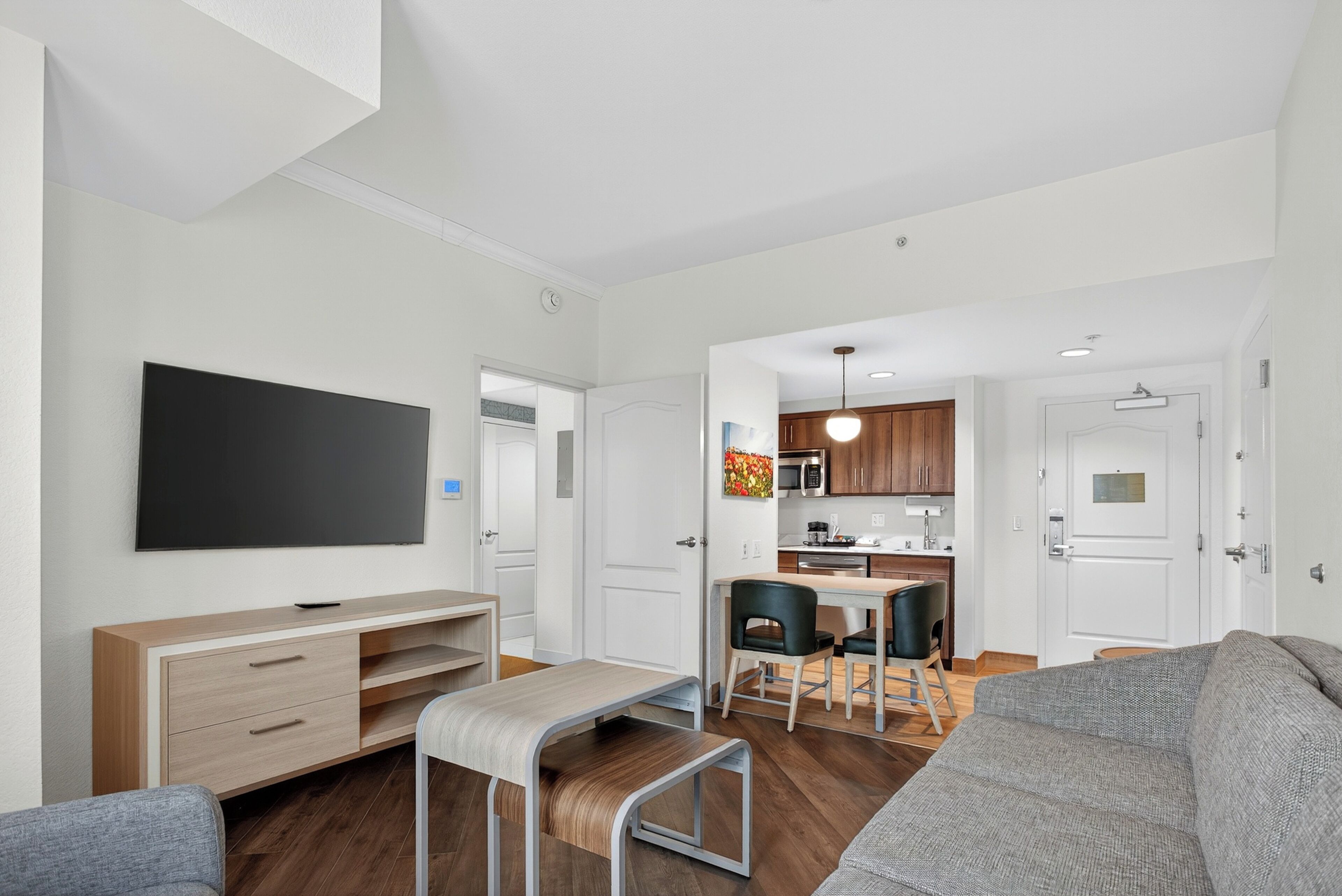 A modern living room with a flat-screen TV, a bench, a sofa, a dining area with a kitchen in the background.