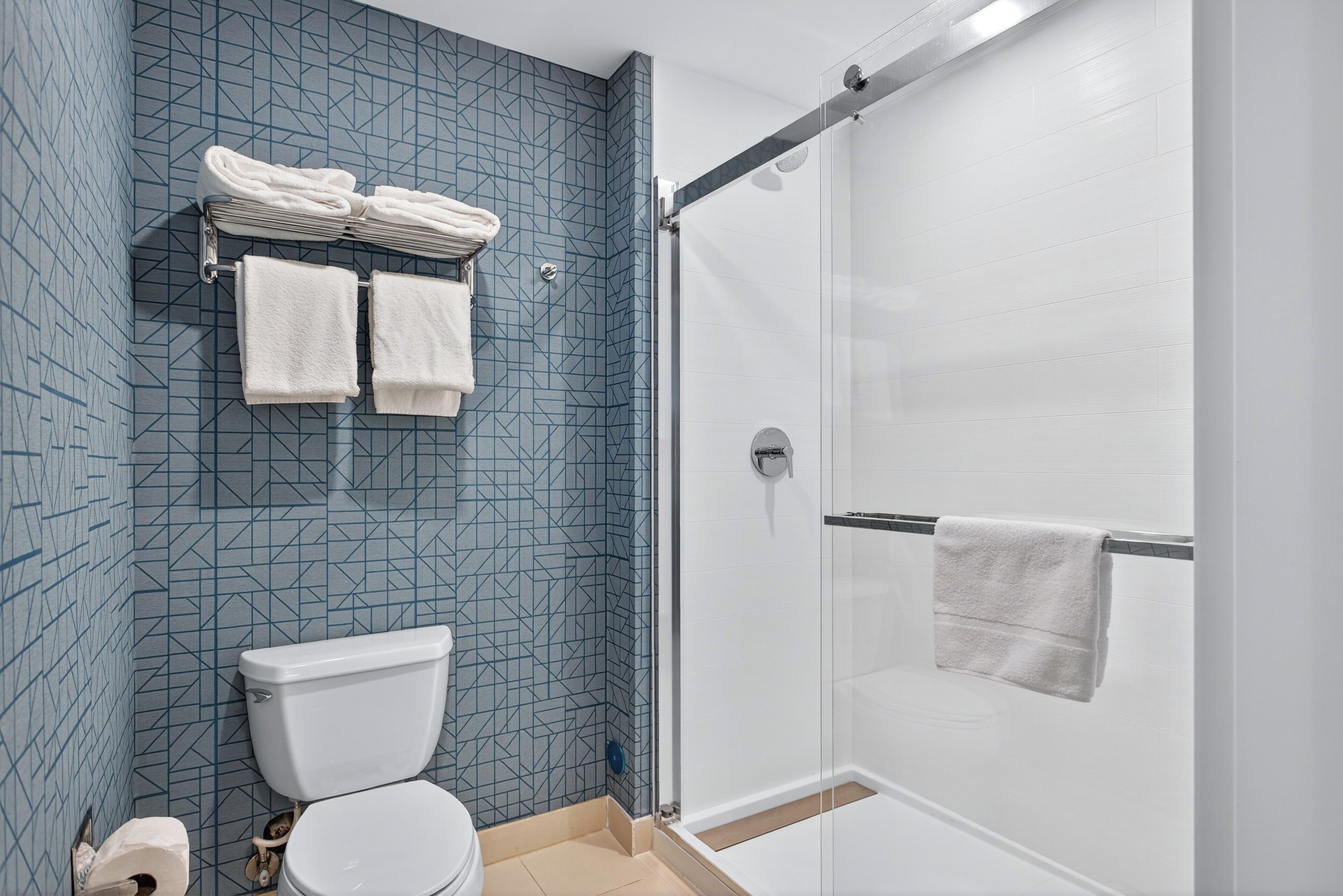 Un baño con un respuesto de azulejos azules geométricos, un inodoro blanco, una ducha con mampara de vidrio y un perchero con toallas dobladas.