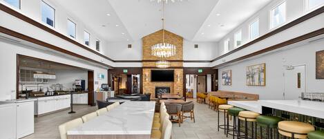 A spacious hotel lobby with a long dining table, bar stools, a fireplace, and a large chandelier.