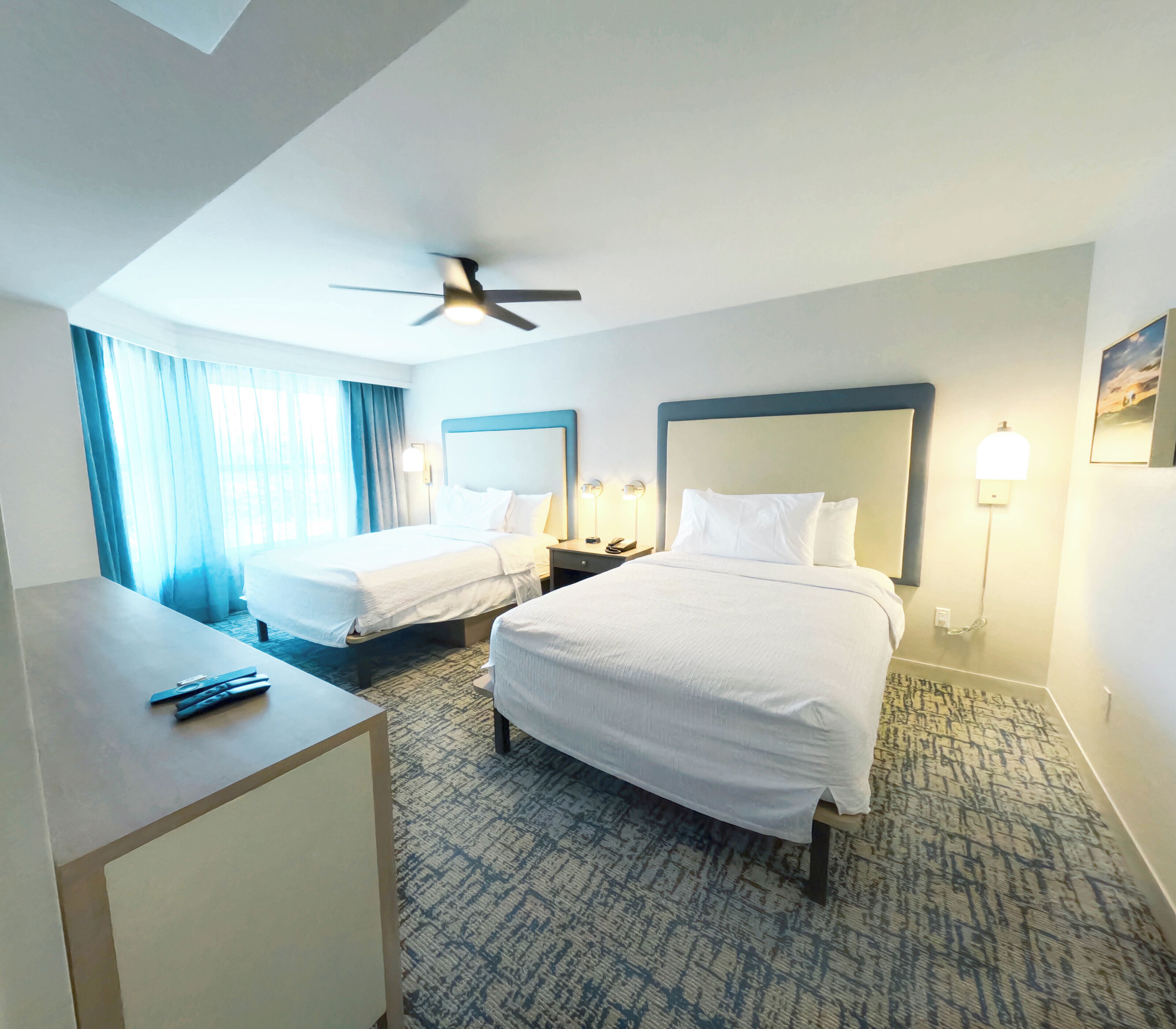 A hotel room with two beds, a desk, a ceiling fan, and a patterned carpet.
