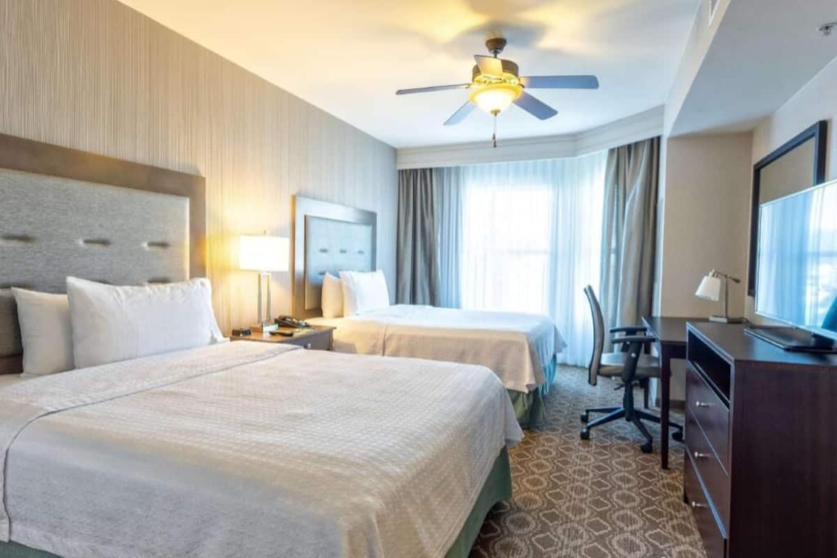 A hotel room with two beds, a desk, a chair, a TV, and a ceiling fan.