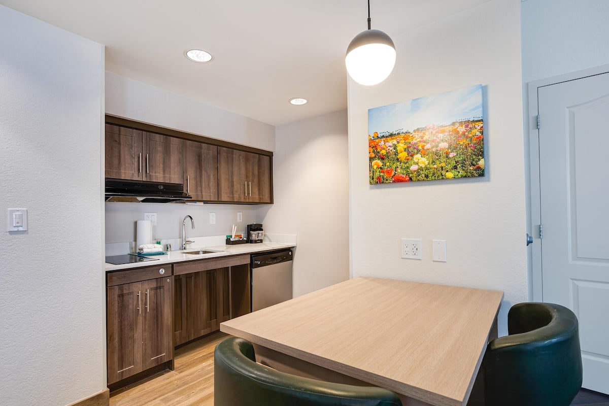 A modern kitchen with wooden cabinets, a stainless steel range hood, a dishwasher, and a light wooden dining table with two chairs.