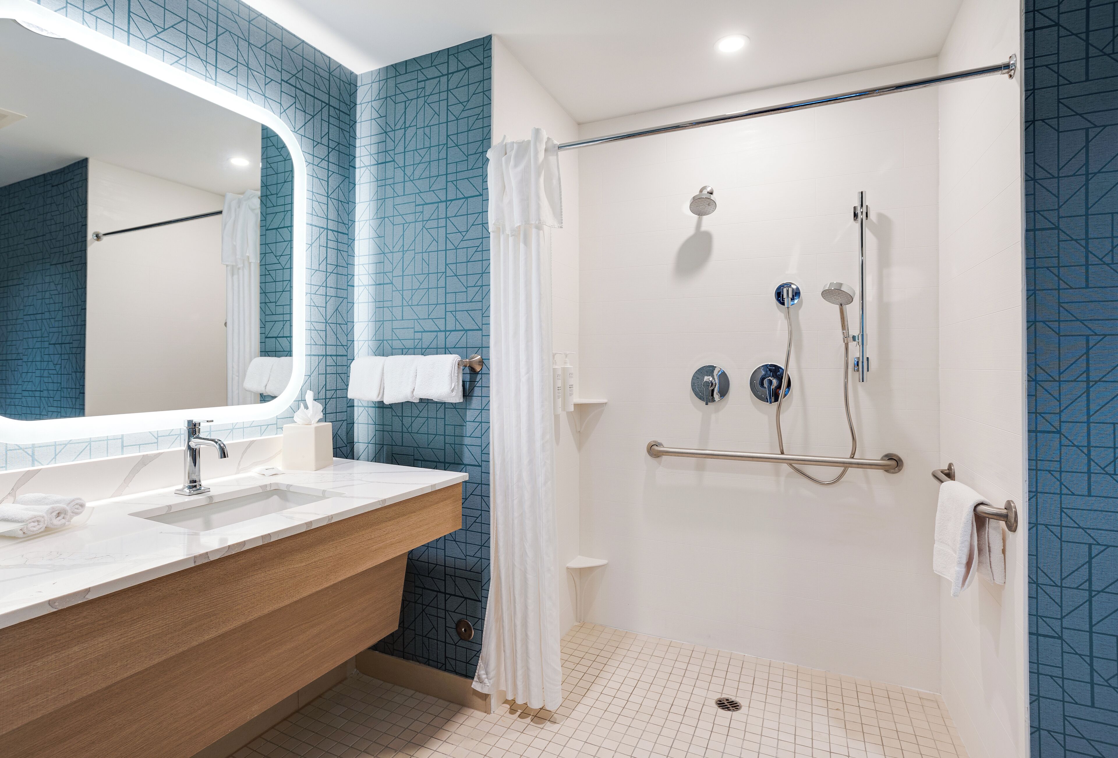A modern bathroom with a large mirror, a sink, a shower with a handrail, and a tiled floor.