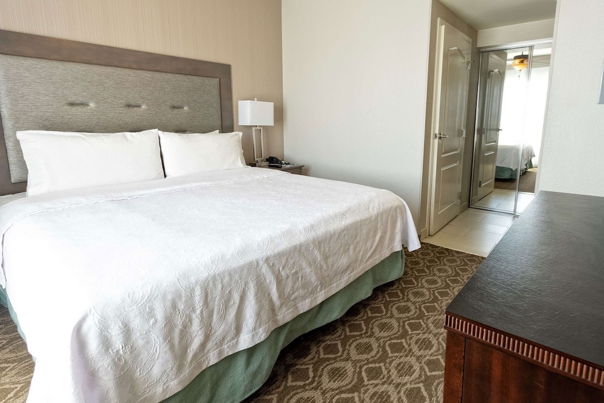 A hotel room with a large bed, a bedside table, a lamp, and a door leading to another room.
