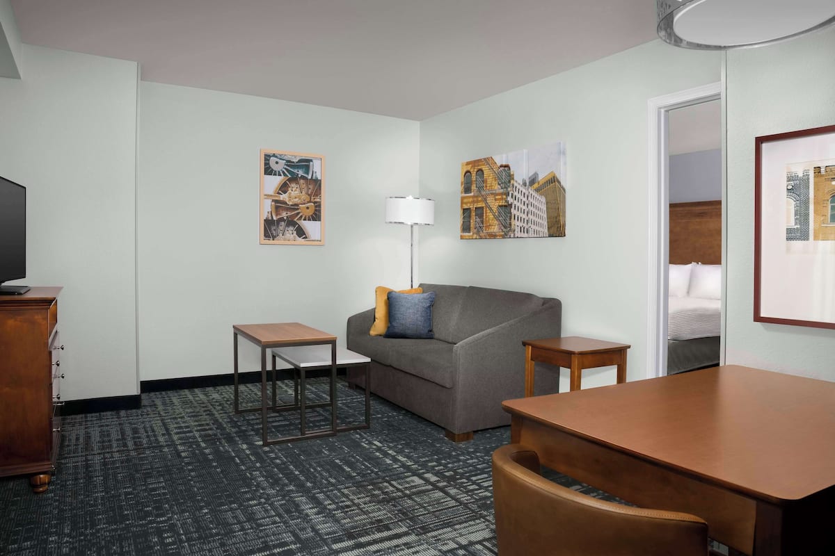 A hotel room with a gray sofa, a small wooden table, a TV on a wooden cabinet, and artwork on the walls.