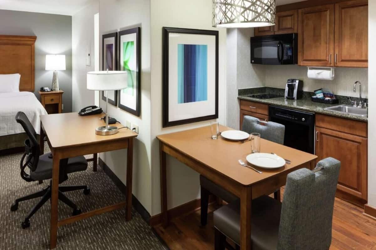 A hotel room with a bed, a desk with a chair, a dining table with chairs, a microwave, a coffee maker, and a sink.