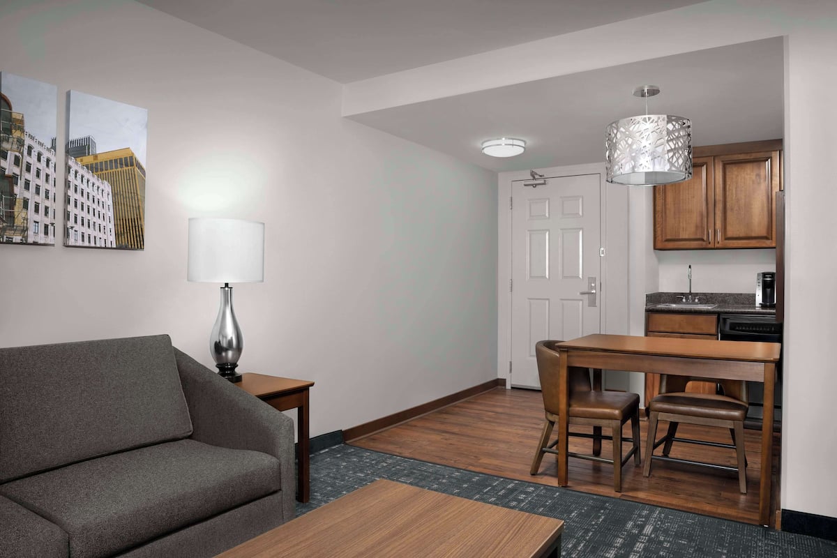 A modern living room with a sofa, a coffee table, and a dining area with a table and chairs. There is a kitchen area with wooden cabinets and a refrigerator.