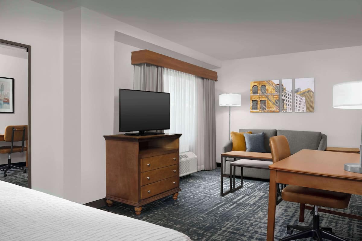 A hotel room with a bed, a desk with a chair, a television on a wooden dresser, a sofa with cushions, and artwork on the wall.