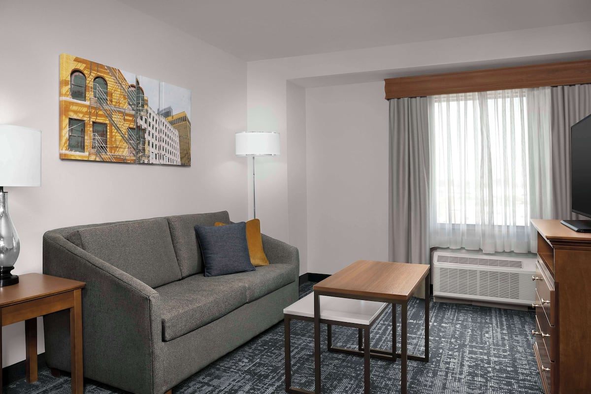 A hotel room with a grey sofa, a wooden side table, a white side table, a small wooden table, a TV, a heater, and a window with curtains.